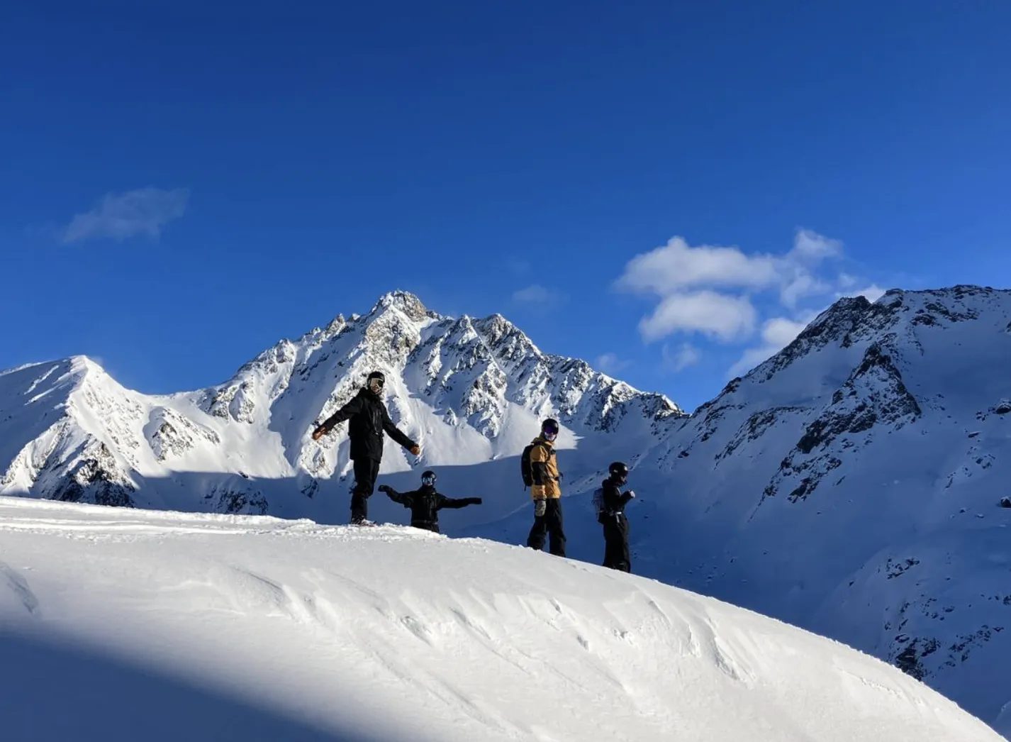 Vier Freerider auf einem verschneiten Grat unter einem felsigen Gipfel bei blauem Himmel