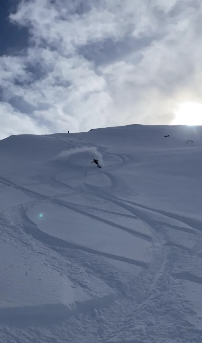 Ein Snowboarder im Schwung im frischen Pulverschnee unterhalb eines Grats, mit älteren Spuren am Hang