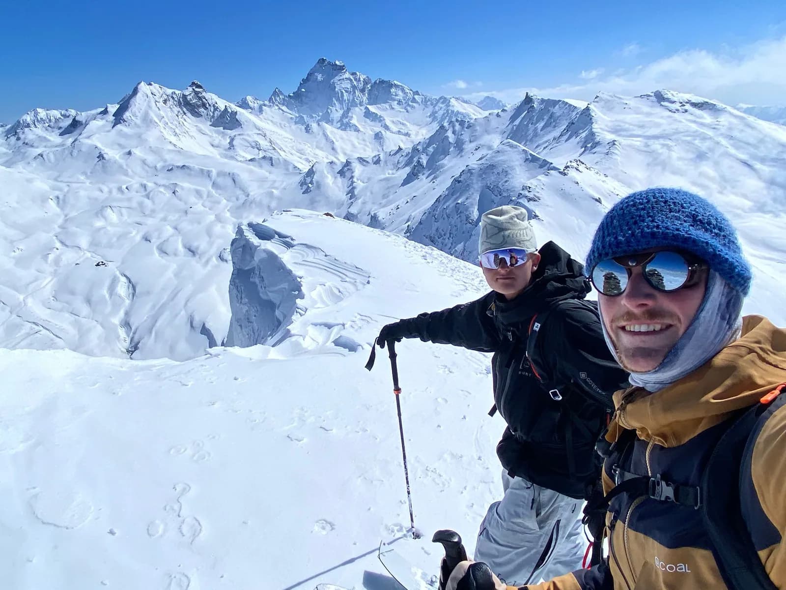Ziggy, vorne mit der blauen Mütze, und Luuk hinter ihm auf einem verschneiten Gipfelgrat mit Stöcken und Splitboards, eine Windlippe unter ihnen und eine Kette von Gipfeln dahinter
