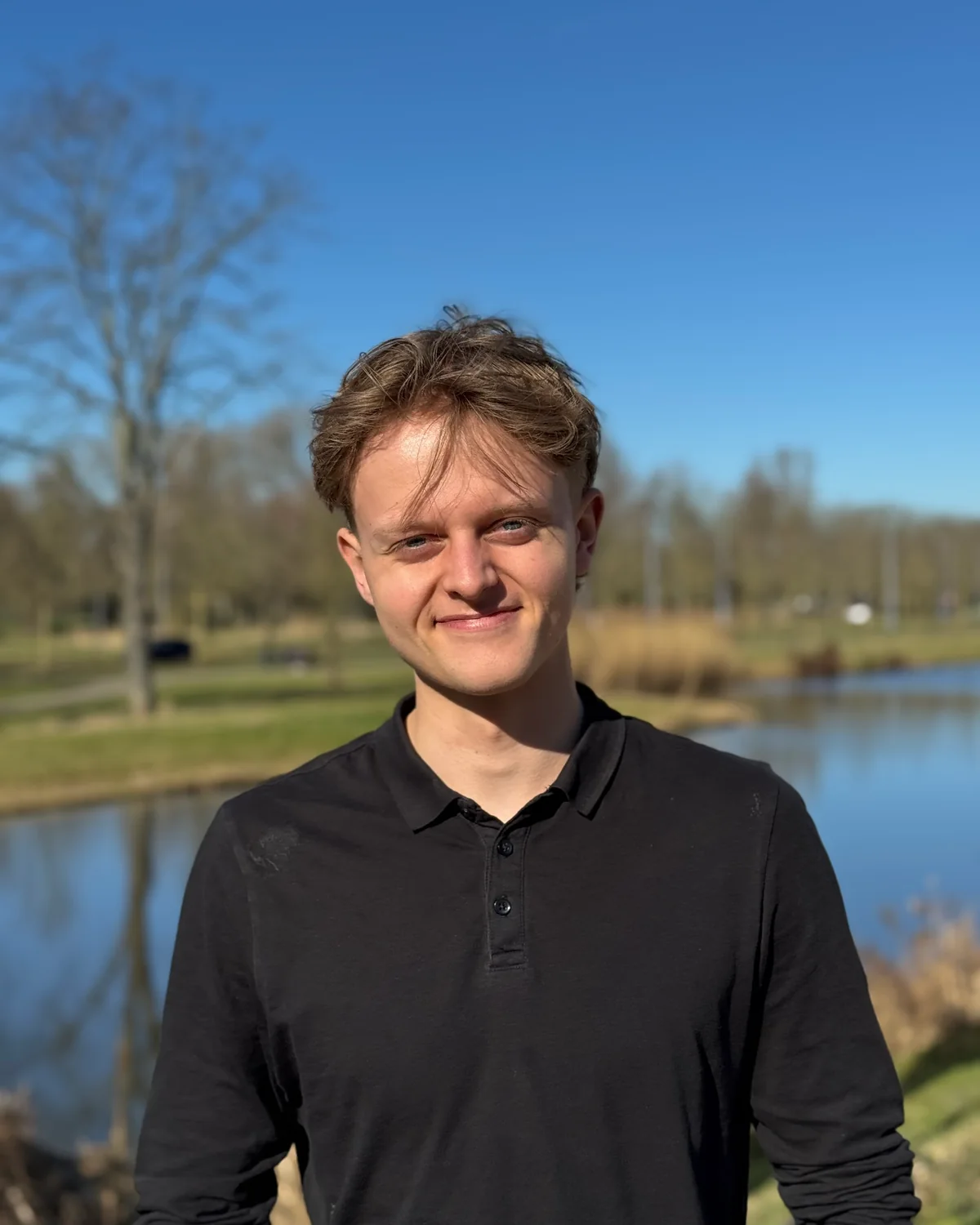 Luuk van der Poel, portrait en extérieur en polo noir, un étang et des arbres nus derrière lui