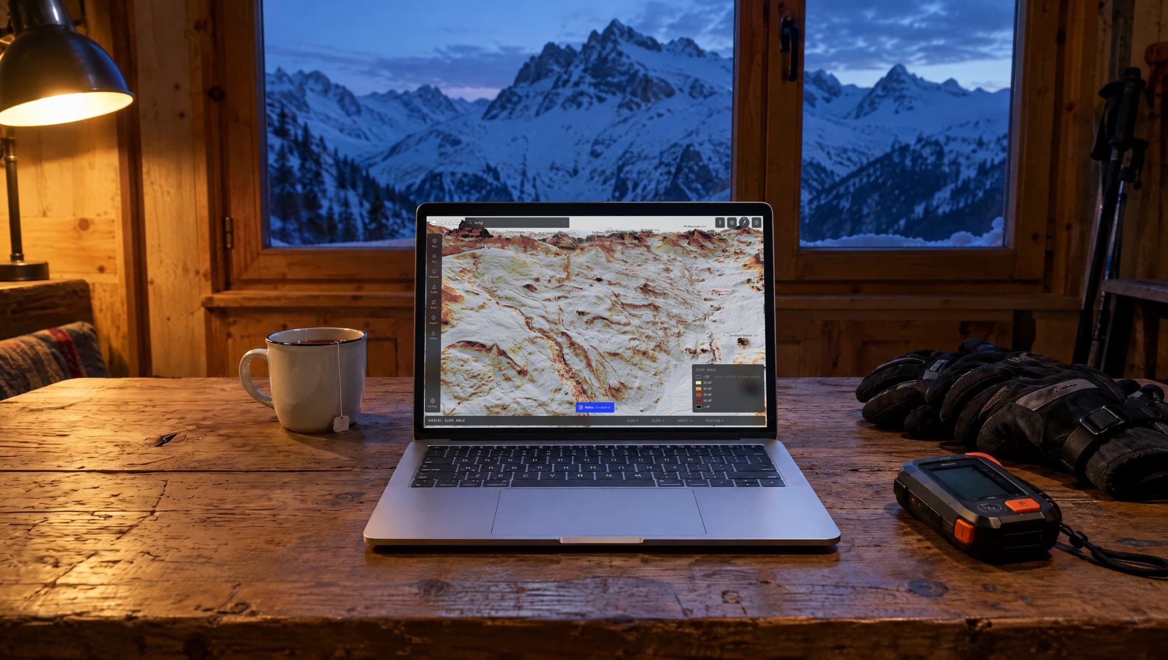 Laptop auf einem Holztisch in einer Berghütte in der Dämmerung, mit der BEYOND-Karte und der Hangneigungsebene rund um die Heidelberger Hütte bei Ischgl, daneben Tee, Handschuhe und ein LVS-Gerät